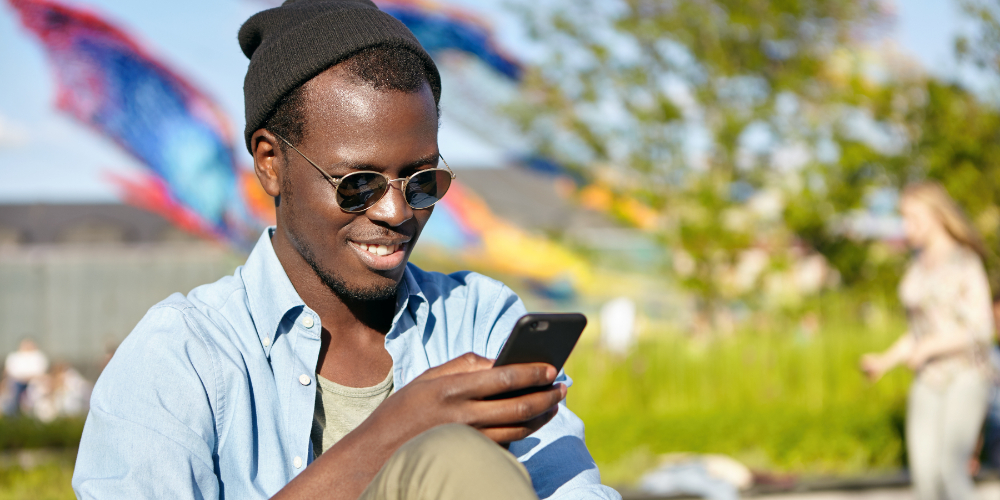 Eleganter dunkelhäutiger Mann mit Dreitagebart sitzt draußen und liest seinen News-Feed oder sendet Nachrichten über soziale Netze auf seinem Smartphone per Gratis-WLAN. Ein glücklicher Hipster, der erfreuliche Neuigkeiten auf seinem Smartphone empfängt.