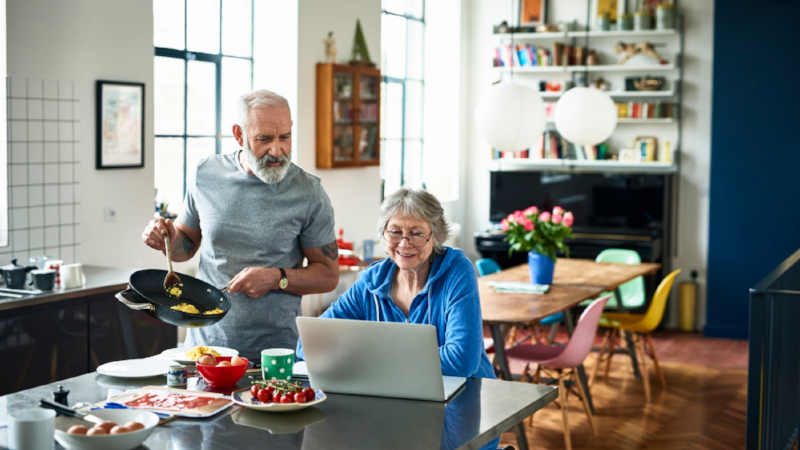 Senior woman using laptop and smiling as man serves breakfast