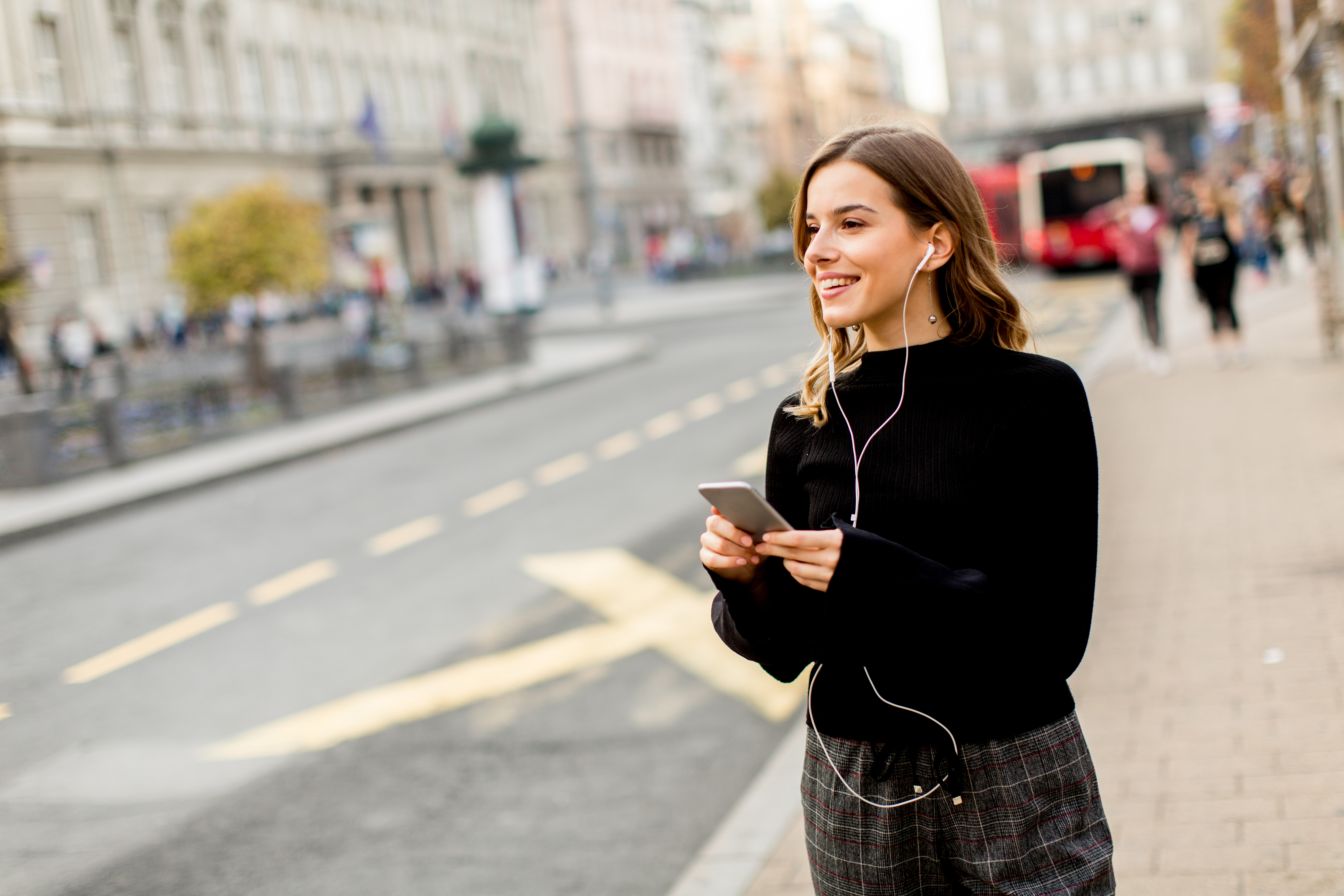 Young brunette woman using mobile while standing on street and waiting for a bus or taxi