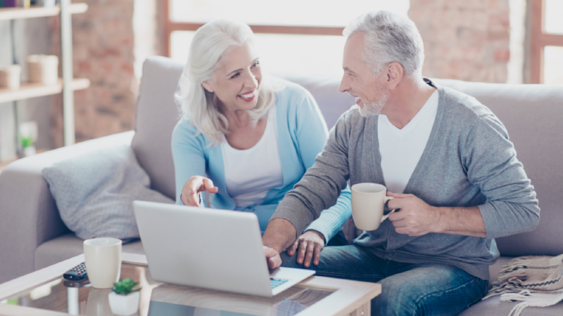Two elderly people are sitting at home at the weekend, drinking coffee and watching films using the computer on the table, the woman is pointing on the monitor