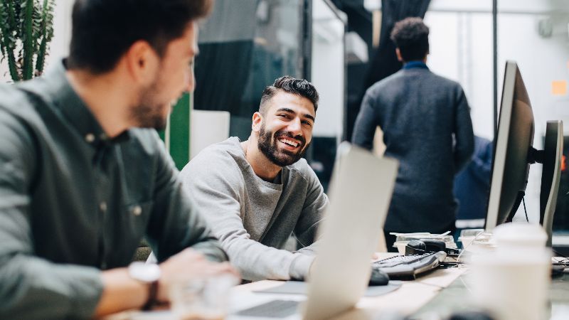 Smiling colleagues looking at each other while sitting by desk in office