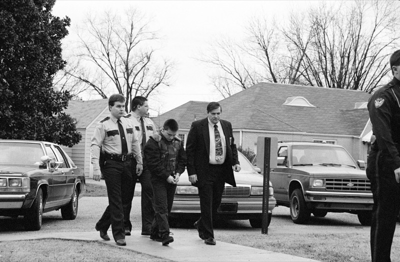 Jessie Misskelley wearing handcuffs being escorted by police officers and a detective