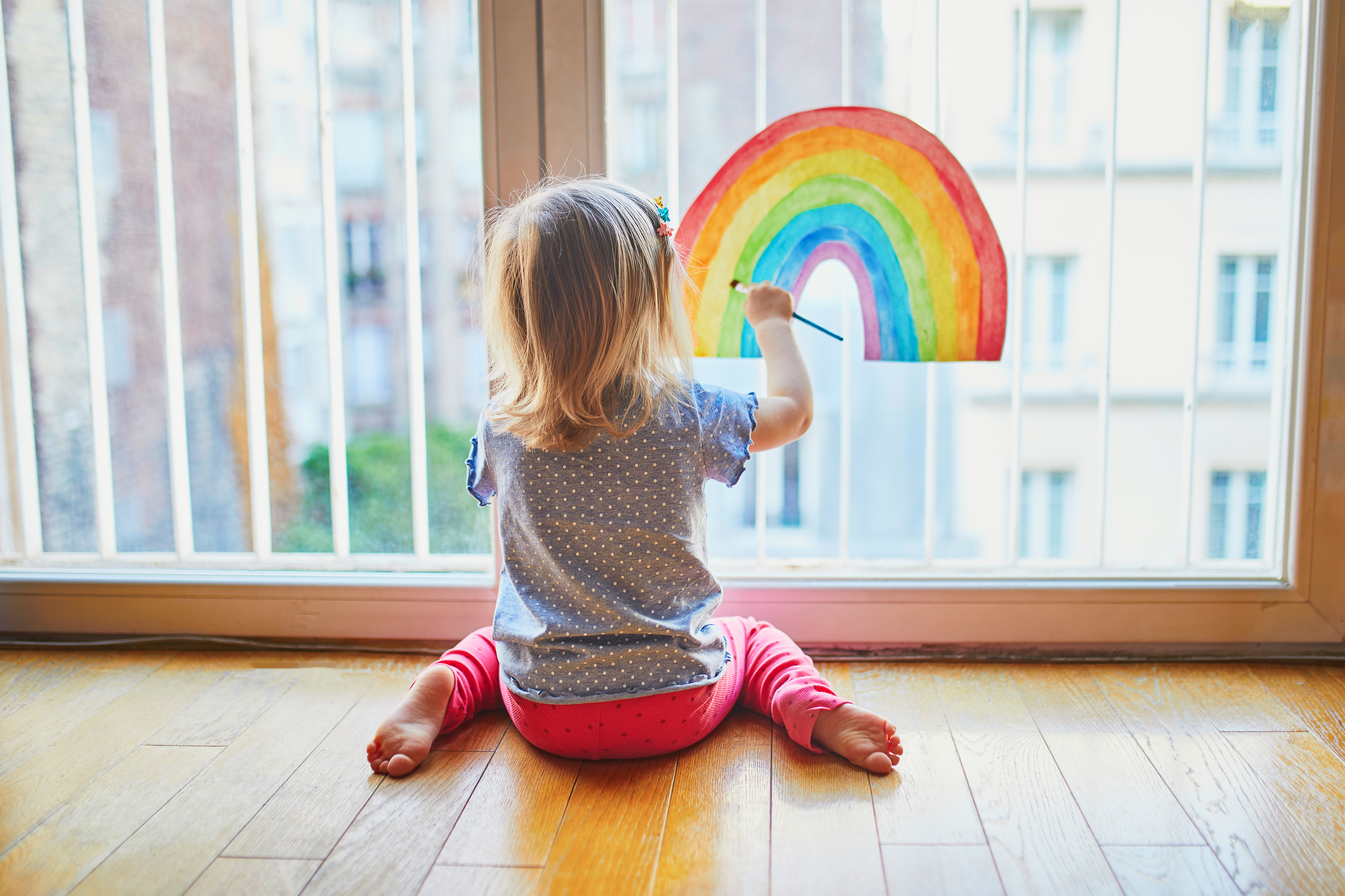 Adorable toddler girl painting rainbow on the window glass