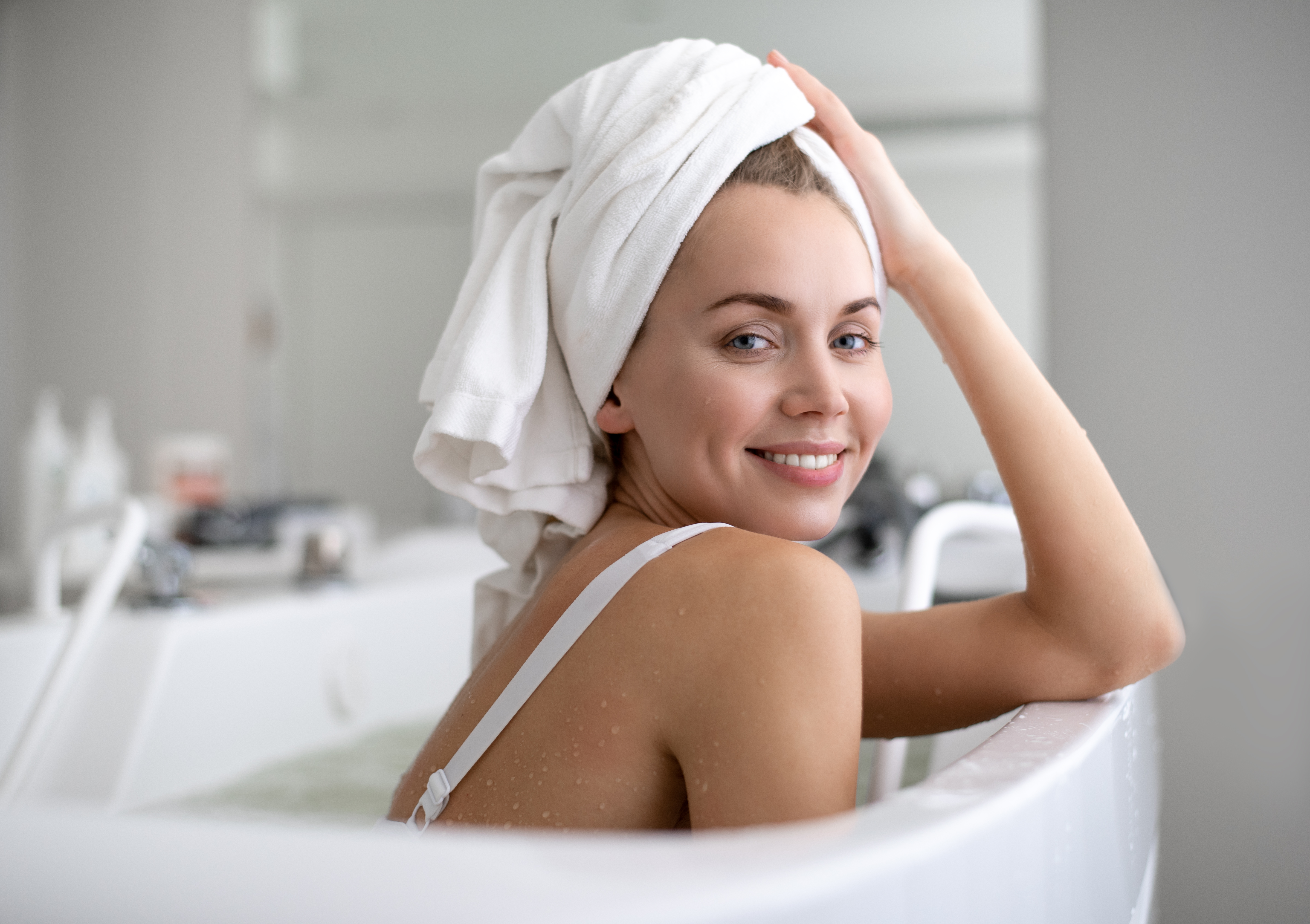 Woman sitting in the bath, smiling, with a white towel wrapped around her head